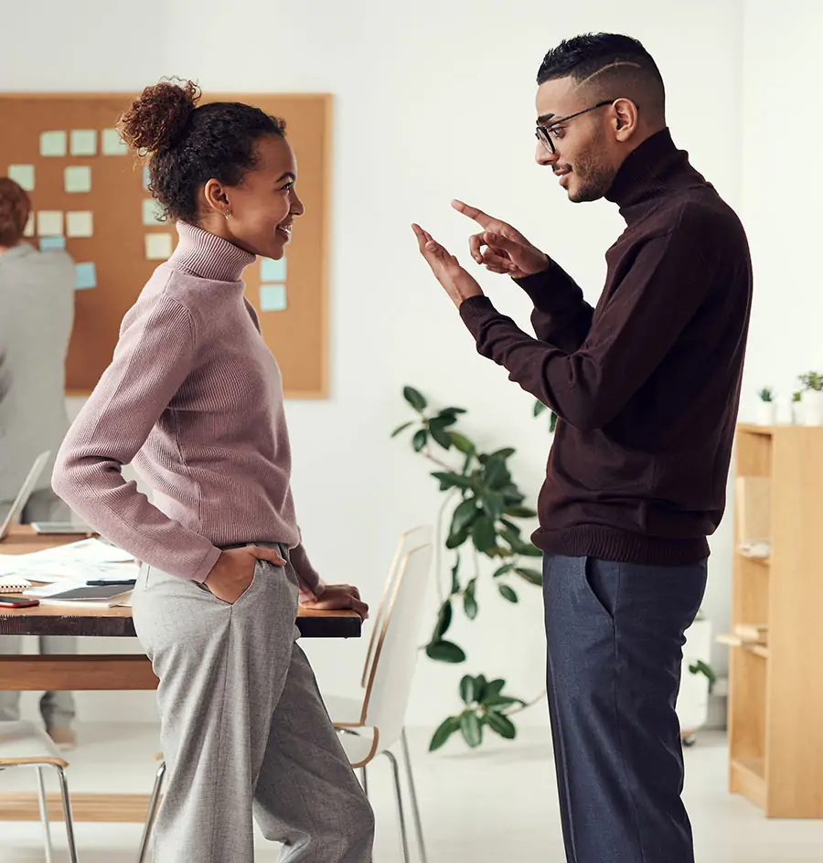 Diverse colleagues discussing a project in a modern office; man in brown turtleneck gesturing while woman listens. Teamwork and professional communication.