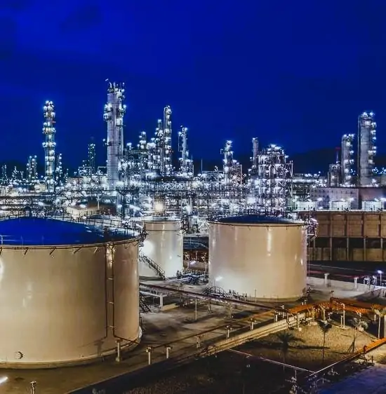 Large industrial oil refinery and petrochemical plant at night, featuring brightly lit distillation towers and large white storage tanks. Energy, chemical processing, and infrastructure industry visual.
