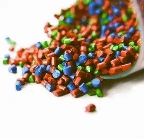 Close-up of multicolored plastic resin pellets (red, blue, and green) spilling out of a clear container onto a white background. Polymer materials, plastic manufacturing, and injection molding concept.